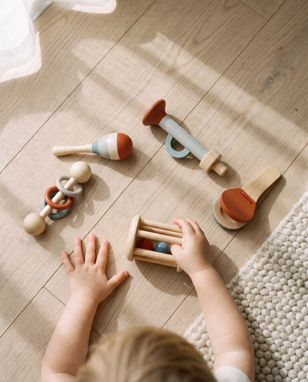 Manos de niño pequeño agitando el rodillo sonajero de Eco, con el resto de instrumentos alrededor sobre suelo de roble
