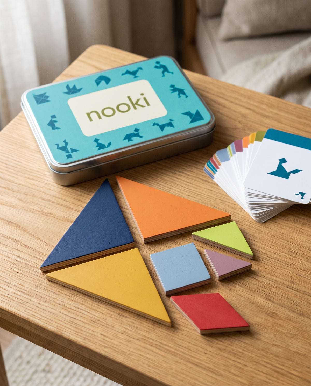 Enigma, tangram de madera de 7 piezas de Nooki, con la caja metálica abierta y el mazo de tarjetas de retos sobre una mesa de roble