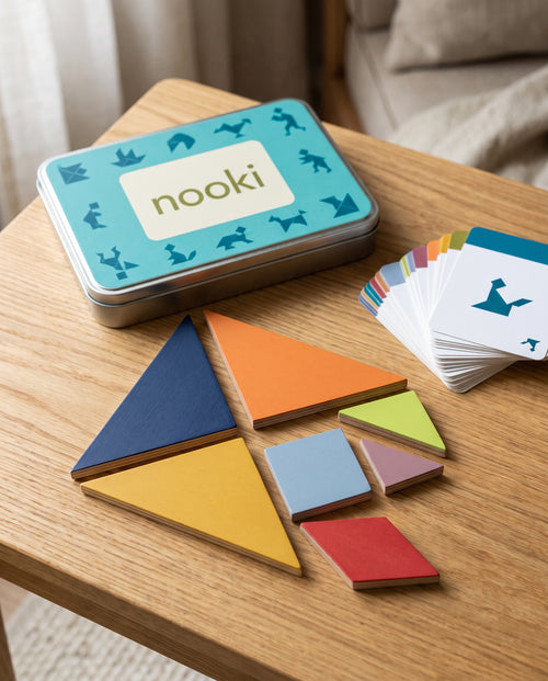 Enigma, tangram de madera de 7 piezas de Nooki, con la caja metálica abierta y el mazo de tarjetas de retos sobre una mesa de roble